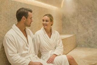 A couple relaxes in a steam room, wearing white robes, enjoying a light conversation in a serene, spa-like atmosphere.