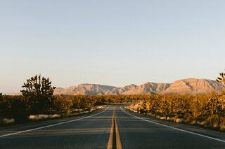 A long, straight road stretches through a desert landscape, framed by rugged mountains and sparse vegetation under a clear sky.