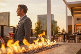 A sophisticated man in a suit stands near a fire pit, holding a drink, while enjoying a sunset view over a city skyline.