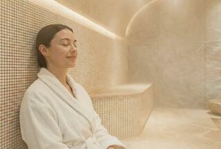 A woman relaxes in a steam room, wearing a white robe with a serene smile, enjoying a peaceful moment in a soothing atmosphere.
