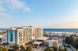 A modern hotel stands near the beach, surrounded by palm trees, with a clear sky and ocean view in the background.