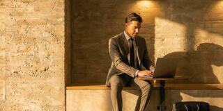 A man in a suit sits on a bench, working intently on a laptop, illuminated by warm lighting against a textured wall.
