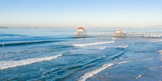 A serene coastal view features gentle waves lapping against a wooden pier with a red-roofed structure under a clear blue sky.