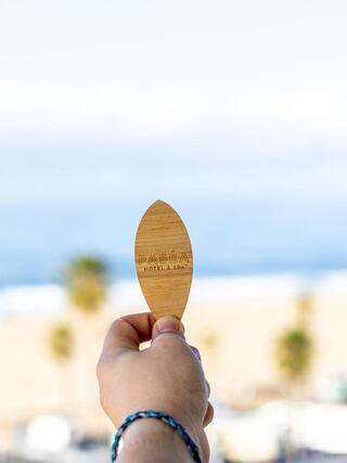 A hand holds a wooden keycard for Paséa Hotel & Spa, with a blurred beach and ocean in the background, evoking a relaxing getaway.