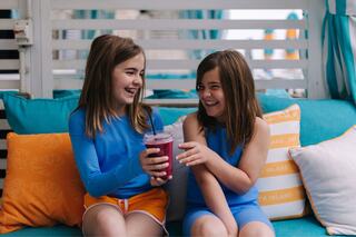 Two smiling girls in colorful outfits share a drink, sitting on a cozy couch with decorative pillows, radiating joy and friendship.
