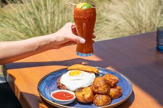 A hand holds a drink next to a plate with a burger topped with a fried egg and golden crispy potatoes, accompanied by a dipping sauce.