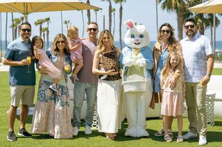 A cheerful group of adults and children pose with a costumed Easter Bunny against a sunny seaside backdrop, enjoying a festive outing.