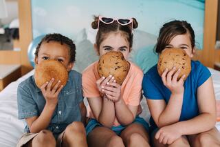 Three children sitting on a bed, holding giant cookies in front of their faces, smiling playfully, dressed in casual summer clothes.