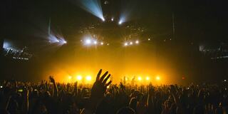 A vibrant concert scene with a sea of hands raised, illuminated by striking golden stage lights against a dark backdrop.
