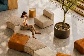 A woman sits comfortably in a modern, airy space, sipping a drink beside stylish, modular seating and a small indoor tree.
