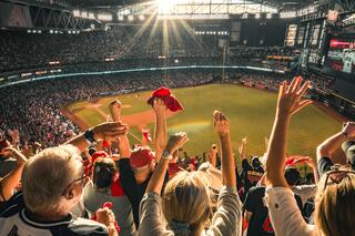 A lively crowd cheers and waves red rally towels in a packed stadium, basking in sunlight as excitement fills the air.