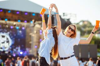 Two young women celebrate at a festival, raising their hands and cups in joy against a vibrant background of a stage and crowd.