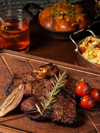 Juicy grilled steak, garnished with rosemary, accompanied by roasted tomatoes, onion, and a cocktail in a warm, inviting setting.