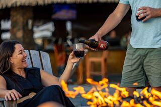 A woman joyfully receives a drink from a man, surrounded by a warm fire, creating a cozy and cheerful atmosphere.
