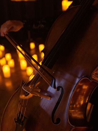 A close-up of a cello being played, illuminated by soft candlelight in the background, creating a warm and intimate atmosphere.
