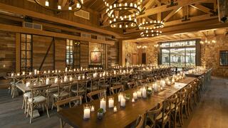 A spacious, rustic dining area with wooden accents, large tables, and glowing candles, illuminated by stylish chandeliers and natural light.