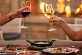 Two people toast with wine glasses beside a delicious spread of gourmet dishes, set against a warm, cozy fireplace backdrop.