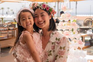 A joyful woman and a young girl share a warm embrace, both wearing floral attire, surrounded by elegant decorations and a shimmering dessert display.