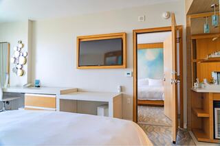 A modern hotel room featuring a bed, desk, TV, and a connecting door to another room, with light decor and stylish furnishings.