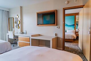 A modern hotel room features two beds, a desk area, a TV, and stylish decor, with light-colored walls and a beach-themed accent.