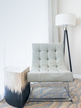 A modern gray tufted chair sits beside a cylindrical wooden side table, complemented by a stylish floor lamp in a bright, airy room.