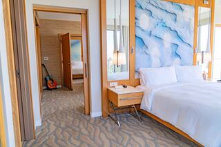 A stylish bedroom features a comfortable bed, modern decor, and a guitar, with light streaming in through large windows.