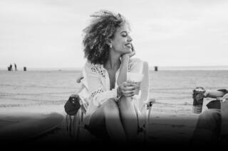 A joyful woman with curly hair smiles while holding a drink, seated on a beach, enjoying a relaxed moment with friends.