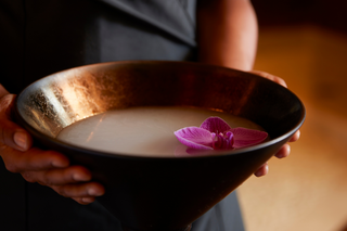 A person holds a deep bowl filled with a milky liquid, adorned with a vibrant orchid, set against a warm, softly blurred background.