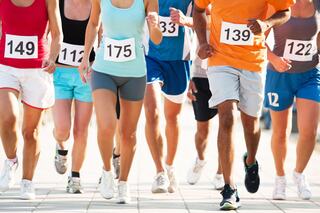 A diverse group of runners strides forward during a race, all wearing numbered bibs on bright athletic gear.
