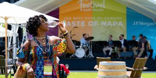 A woman in a colorful dress enjoys a drink at a lively outdoor festival, with a band performing on stage in the background.
