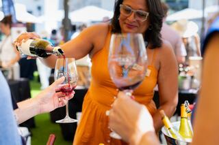 A woman in an orange dress pours red wine into a glass, surrounded by a lively outdoor event with wine bottles and eager attendees.