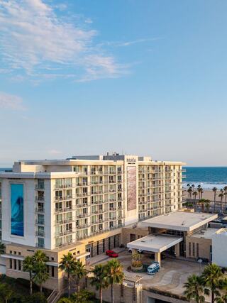 A modern hotel stands near the ocean, featuring a multi-story facade, palm trees, and a welcoming entrance under a clear blue sky.
