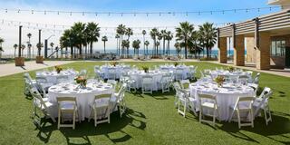 An elegant outdoor event space features round tables with white cloths and floral centerpieces, surrounded by palm trees and a beachfront view.