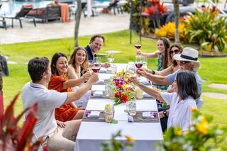 A group of friends cheers with drinks at a vibrant outdoor table, surrounded by lush greenery and colorful flowers, celebrating together.