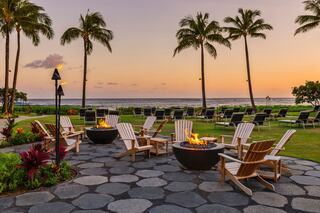 A serene beachfront setting with palm trees, cozy seating around fire pits, and a colorful sunset reflecting on calm waters.