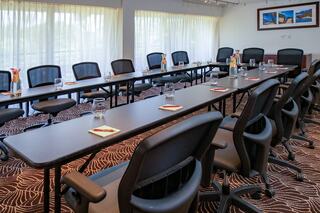 A bright conference room featuring long tables with chairs, glasses of water, coasters, and large windows with sheer curtains.