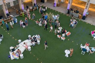 A lively outdoor gathering features guests mingling on green grass, seated in various arrangements amid string lights and a festive atmosphere.