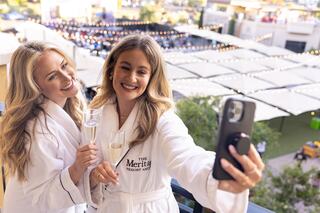 Two women in cozy robes celebrate with champagne while taking a selfie on a balcony, enjoying a festive atmosphere.