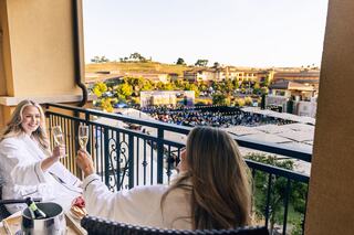 Two women in bathrobes toast with champagne on a balcony, overlooking a lively outdoor concert, surrounded by scenic vineyards.