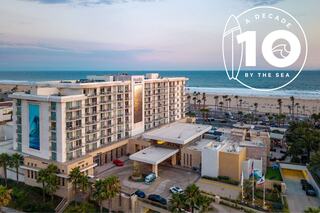 Two surfers ride gentle waves in front of a beachfront hotel, celebrating a decade of seaside experiences. Sunlit palms enhance the vibe.