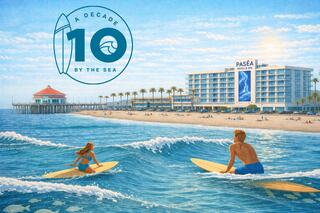 Two surfers ride gentle waves in front of a beachfront hotel, celebrating a decade of seaside experiences. Sunlit palms enhance the vibe.